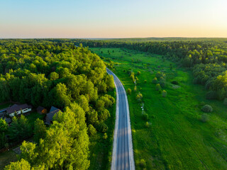 An aerial view shows a winding road cutting through a vast expanse of lush green forest and open fields under a clear sky with a sunset glow