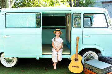 Girl in a straw of a hat and overalls sit near on the doorstep of the motor home. Smile child girl near old hippie bus in Campsite with guitar, suitcases. Concept vacation, trip by car, campervan