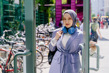 Obraz premium Young Muslim woman exiting building with headphones and backpack in city