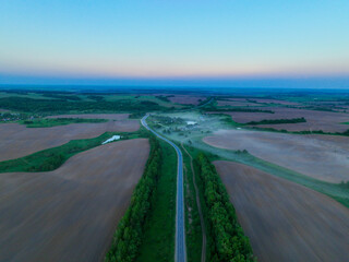 An aerial view captures a long, straight road cutting through expansive misty fields, leading towards distant villages and forests under a soft morning sky