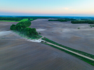 Fototapeta premium An aerial view shows mist covering vast fields, with a rural road and a vehicle visible, leading into a cluster of trees at dawn
