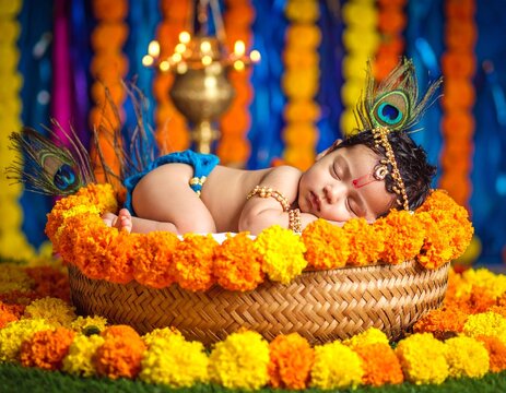 An infant dressed as Lord Krishna sleeping in a hand-woven jhula (cradle) decorated with marigold flowers and peacock feathers. Background includes oil lamps and colorful fabric drapes.