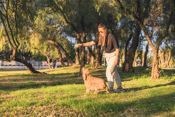 A young woman with long brown hair interacts with a golden cocker spaniel in a sunny park. The dog...