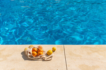 Mesh cotton bag filled with fresh lemons and oranges resting on the edge of a swimming pool under bright sunlight, symbolizing healthy summer lifestyle, eco-friendly living