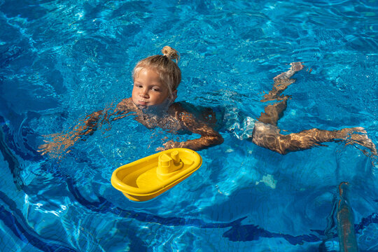 Little blond boy swimming underwater in a bright blue pool, holding his breath with puffed cheeks while playing with a floating yellow toy boat on a sunny summer day
