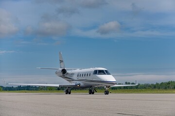 A business jet taxis across the runway under a wide blue sky with distant clouds. Captured from a front quarter angle, the composition showcases both engines, landing gear, and fuselage.