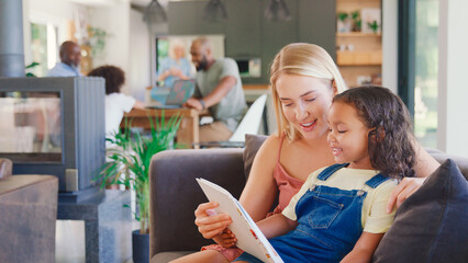 Mother And Daughter Reading Book At Home Together With Multi-Generation Family In Background