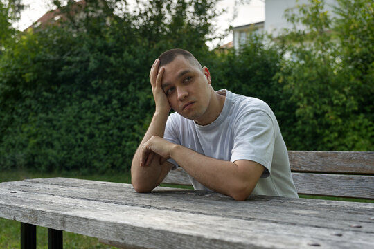 A man with a buzz cut sits at a wooden picnic table outdoors, looking pensive with his hand on his head.
