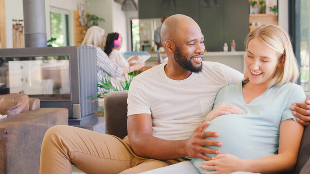 Multi-Racial Couple With Pregnant Woman On Sofa At Home With Multi-Generation Family In Background