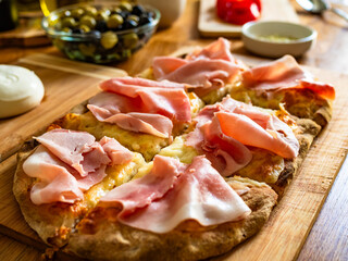 Cooking Pinsa Romana with mozzarella and burrata cheese, boiled ham, tomatoes and olives on wooden kitchen table, close-up