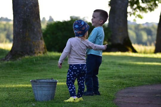 Two toddlers standing on green lawn near metal bucket, one girl with patterned hat reaching arm toward boy. Casual summer clothes, soft natural light, friendly outdoor moment. - Powered by Adobe