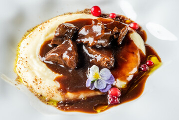 Gourmet beef in rich brown sauce served on mashed potatoes, garnished with edible flower and red berries, presented on a white plate in restaurant style.