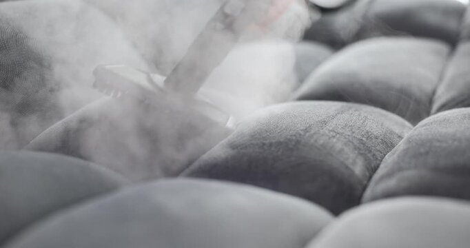 Handheld device emits steam onto tufted gray sofa with moisture and vapor. Process of steam cleaning highlights deep cleaning and sanitization