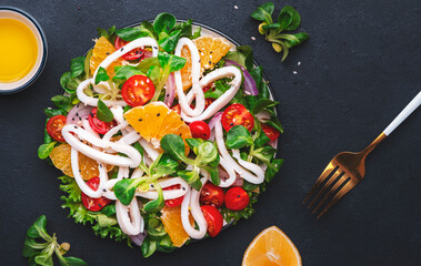 Yummy seafood salad with oranges, squid, tomatoes, lettuce and lemon dressing, black table, top view