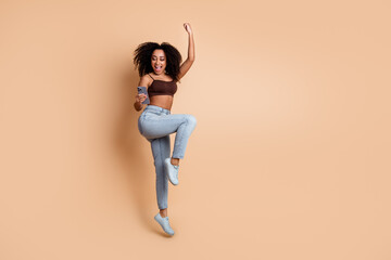 Young cheerful woman with curly hair dancing joyfully while holding a smartphone in a beige...