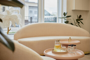 Modern waiting area featuring curved beige sofa and round tables holding glass pitcher with lemon water, two water bottles, small perfume bottle, large window in background