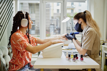 Young biracial woman sitting at manicure table receiving nail treatment from Caucasian female manicurist wearing gloves and face mask, nail polish bottles arranged on table