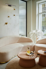 Modern waiting area in beauty salon featuring curved beige sofa, round coffee tables with glass pitcher of water and small bottles, decorative wall art and large window providing natural light