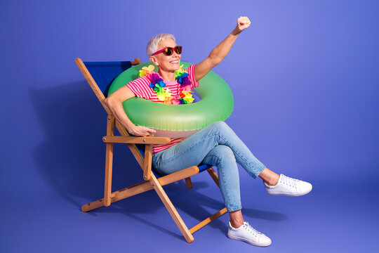 Cheerful mature woman enjoying leisure time wearing fun summer accessories in a chair against colorful background