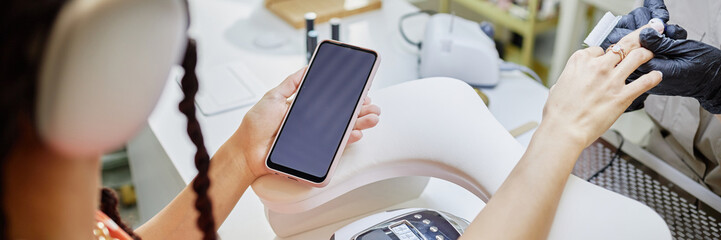 Website header shot of young adult woman holding smartphone while receiving manicure from female professional wearing gloves, closeup of hands in beauty salon, modern nail care procedure in progress