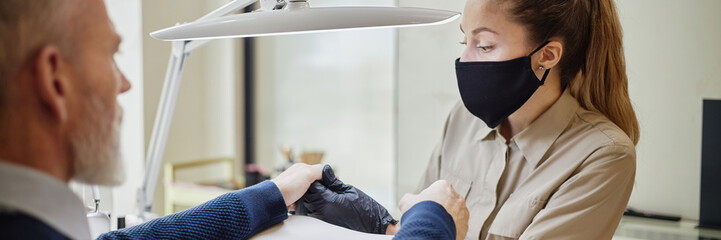 Website header shot of young adult Caucasian woman wearing protective mask performing manicure for middle aged man in salon, both focusing on hands under bright lamp, close up interaction