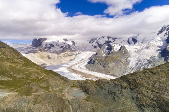 Aerial view of the vast Gorner Glacier snakes through rugged terrain under a bright sky, cradled by the majestic peaks of the Swiss Alps, Gornergrat, Zermatt, Switzerland.