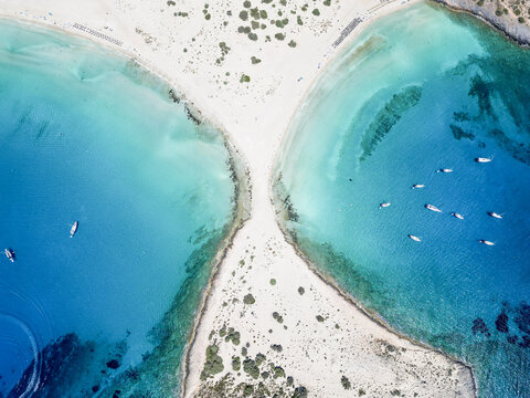 Aerial view of turquoise waters embracing the soft, white sands of a double cove where small boats rest peacefully, Elafonisos, Greece.