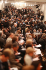 Blurred background image of a crowded press conference room filled with journalists seated and standing, with notepads, cameras, and microphones, all focused on a speaker. News event concept.