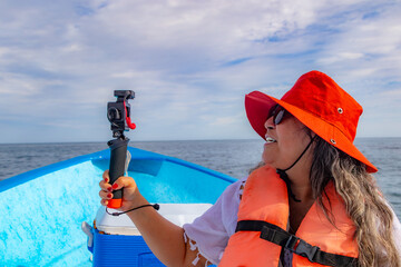 Smiling female tourist filming sea with camera on boat, ocean with horizon in background, sunglasses, hat, and orange life jacket, sunny day in Loreto, Baja California Sur, Mexico