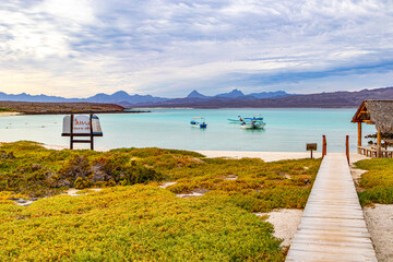 Beach in protected area of Coronado Island, boats anchored on waters of bay, wooden walkway and palm-thatched palapa, mountains in background, National Marine Park, Baja California Sur, Mexico