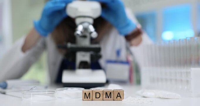 Wooden blocks with MDMA on desk with scientist in blue gloves looks through microscope near test tubes. Forensic science and drug analysis in lab