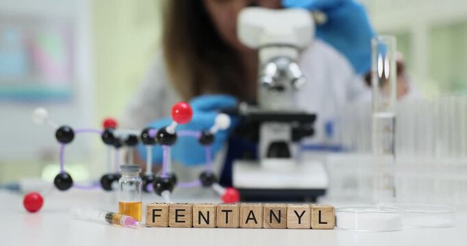 Female scientist looks through microscope near Fentanyl on blocks and molecular model on table. Forensic analysis related to opioid fentanyl