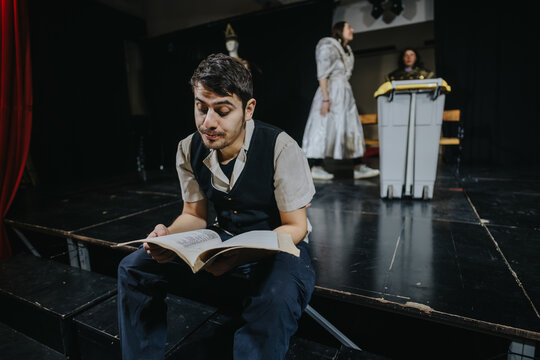 A young man engrossed in reading a book while sitting on a theatre stage, with performers in costumes rehearsing in the background under dramatic lighting.