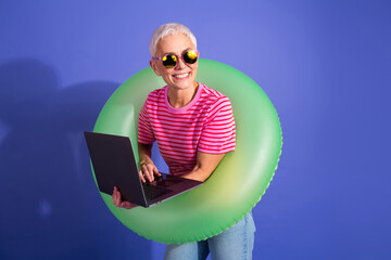 Cheerful senior woman using a laptop and wearing a green inflatable ring against a vibrant purple...