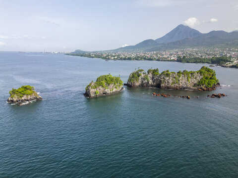 Aerial view of dramatic, rocky islets crowned with vibrant green vegetation, contrasting against the deep blue ocean, with the city and mountain backdrop, Limbe, Southwest Region, Cameroon.
