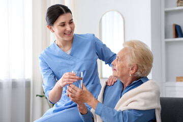 Fototapeta premium Nurse giving glass of water to senior woman indoors. Home health care service