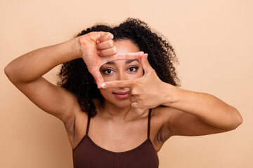 Fototapeta premium Young woman framing her face with fingers against beige background, showing confidence and happiness, trendy casual look