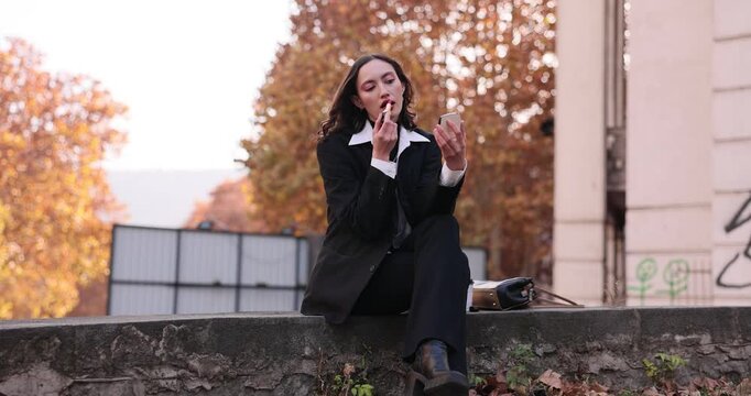 Woman in black suit sits on low parapet applying lipstick while looking into compact mirror. Moment of personal grooming against autumn trees in fall