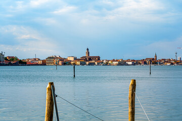  Scenic view of a tranquil waterfron Chioggia, Italy  with wooden posts in foreground, showcasing charming buildings and a church steeple in the distance, ideal for travel inspiration