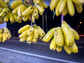 Hanging bunches of ripe yellow bananas are prominently displayed against a blurry, dark night street, likely from a roadside stall, illuminated by artificial light.