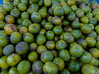 A close-up, high-angle shot of a large, full pile of green and yellow-hued oranges, some with blemishes, creating a dense and textured display of fresh fruit at a market.