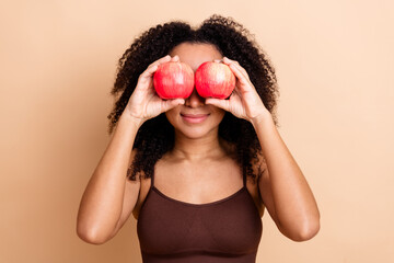 Playful woman with curly hair holds red apples against her eyes, enjoying fun moments on a beige...
