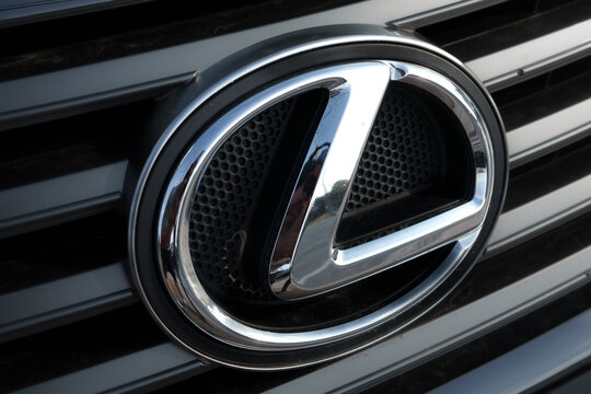 Close-up of a Lexus emblem on a dark grille, showing the chrome L inside an oval with a mesh background.