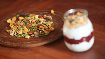 closeup of glass jar filled with creamy yogurt and a layered mix of crunchy granola and muesli, placed on rustic wooden table with scattered nuts and seeds on wooden plate.