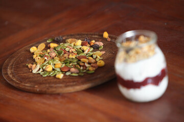 closeup of glass jar filled with creamy yogurt and a layered mix of crunchy granola and muesli, placed on rustic wooden table with scattered nuts and seeds on wooden plate.