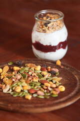 closeup of glass jar filled with creamy yogurt and a layered mix of crunchy granola and muesli, placed on rustic wooden table with scattered nuts and seeds on wooden plate.
