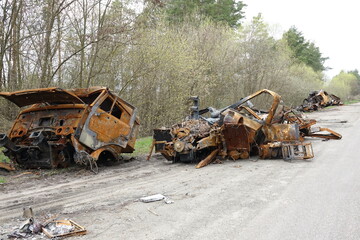 Abandoned burnt vehicles with rusted frames and damaged parts left on a forest roadside. Destroyed machinery shows signs of fire and structural impact in an outdoor rural environment.