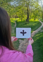Woman hands holding a paper with arrows pointing left, right, and straight directions white standing in front of a crossroad. Forked path symbol for decision making and life choices concept