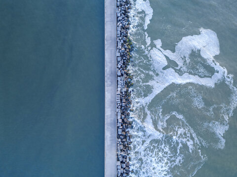 Aerial view of a stark concrete pier dividing the tranquil, deep blue sea from the frothy, turbulent waves crashing against the stone breakwater, Wijk aan Zee, North Holland, Netherlands.