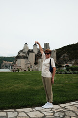 Fototapeta premium Woman posing with raised hand pretending to hold a tower of Golubac Fortress in a playful travel photo. Travel in Serbia concept